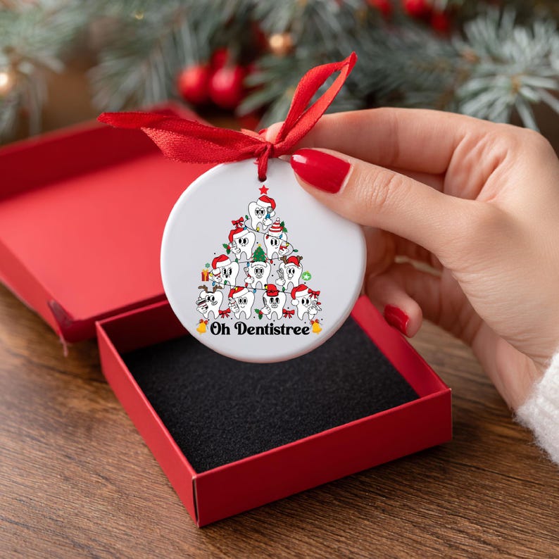 Puede incluir: Un adorno navide&ntilde;o de cer&aacute;mica blanca con una cinta roja, con un dise&ntilde;o festivo de dientes dispuestos como un &aacute;rbol de Navidad. El adorno est&aacute; en una caja de regalo roja, con el texto "Oh Dentistree".