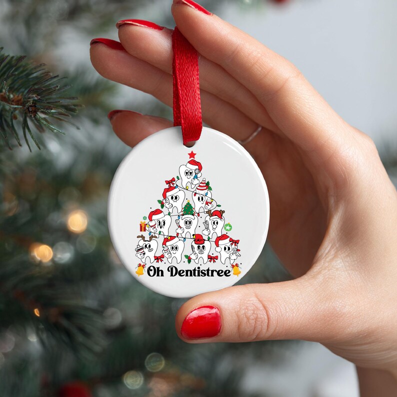 Puede incluir: Un adorno navide&ntilde;o redondo y blanco con una cinta roja. El adorno presenta un &aacute;rbol de Navidad de dibujos animados hecho de dientes sonrientes con gorros de Pap&aacute; Noel. El texto "Oh Dentistree" est&aacute; en la parte inferior.