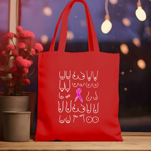 May include: Red tote bag with a white design of various breast shapes and a pink ribbon. The text "You are strong" is written at the bottom of the bag.