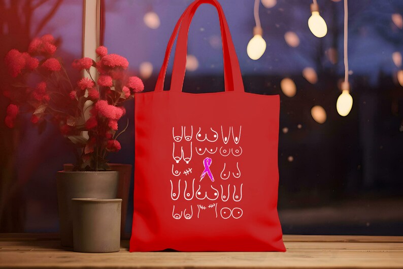 May include: Red tote bag with a white design of various breast shapes and a pink ribbon. The text "You are strong" is written at the bottom of the bag.