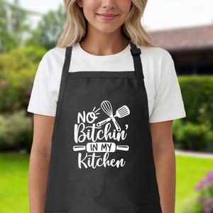 May include: Black kitchen apron with white text that reads "No Bitchin' in my Kitchen" and a graphic of a whisk and spatula.
