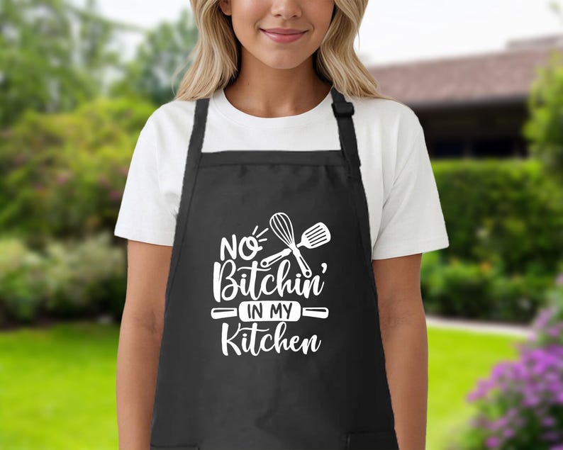 May include: Black kitchen apron with white text that reads "No Bitchin' in my Kitchen" and a graphic of a whisk and spatula.