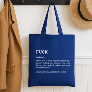 May include: A navy blue tote bag with white text that reads "FUCK" followed by a definition. The bag has long straps and is hanging on a wall. A tan coat and a straw hat are also visible.