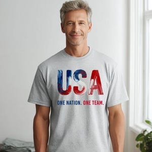 K&ouml;nnte beinhalten: Ein graues T-Shirt mit dem Wort "USA" in rot, wei&szlig; und blau. Darunter steht der Text "ONE NATION. ONE TEAM."