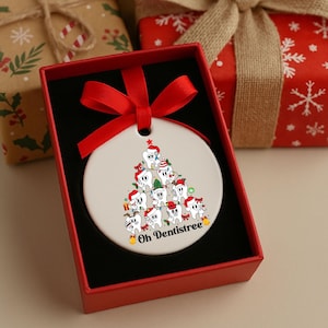 Puede incluir: Adorno navide&ntilde;o de cer&aacute;mica blanca en una caja roja. El adorno presenta un dise&ntilde;o de &aacute;rbol de Navidad hecho de dientes de dibujos animados con gorros de Pap&aacute; Noel. El texto "Oh Dentistree" est&aacute; impreso en la parte inferior.