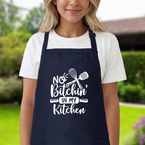 May include: A navy blue kitchen apron with white text that reads "No Bitchin' in my Kitchen" and a white silhouette of a whisk and spatula.