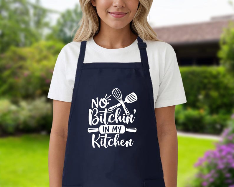 May include: A navy blue kitchen apron with white text that reads "No Bitchin' in my Kitchen" and a white silhouette of a whisk and spatula.