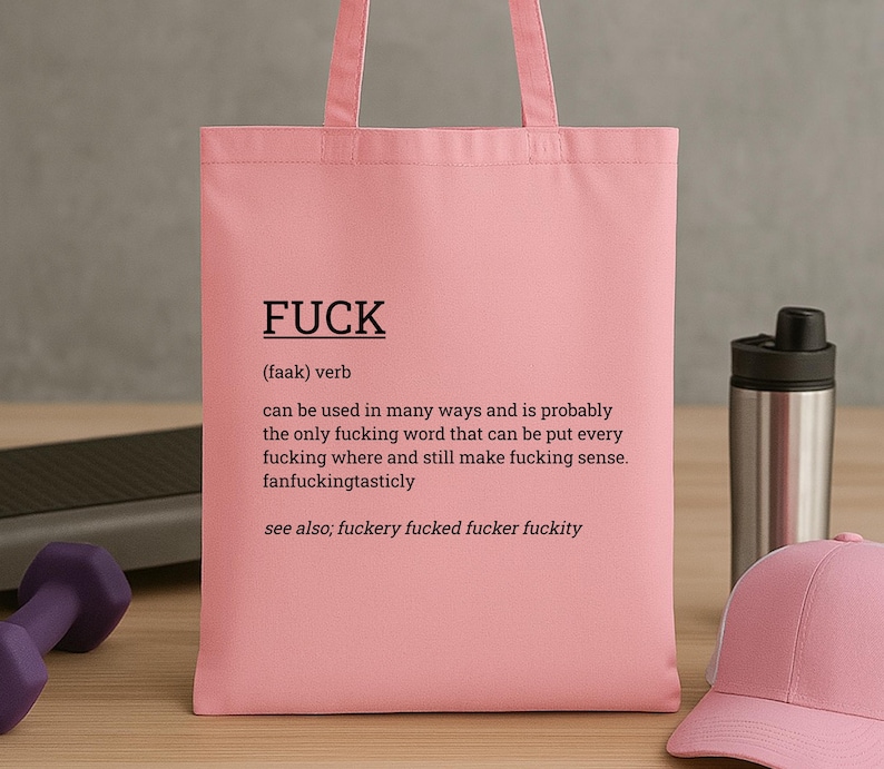 May include: A pink tote bag with the word "FUCK" printed in black, along with a definition. The bag is made of a soft material and has handles for carrying. A pink baseball cap and a water bottle are in the background.