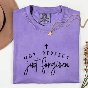 May include: A lavender Comfort Colors t-shirt with the text "NOT PERFECT just forgiven" in black script. A gold necklace with a star pendant rests on the shirt. A brown hat is visible in the upper left corner.