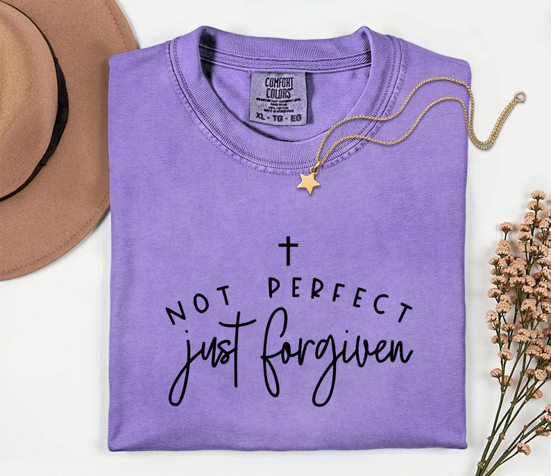 May include: A lavender Comfort Colors t-shirt with the text "NOT PERFECT just forgiven" in black script. A gold necklace with a star pendant rests on the shirt. A brown hat is visible in the upper left corner.