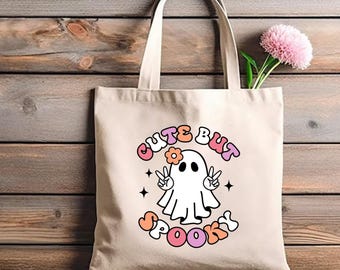 May include: A beige canvas tote bag featuring a graphic of a white ghost making peace signs with its hands. The ghost wears a small orange flower on its head. The text "CUTE BUT SPOOKY" is printed in a curved, multi-colored font around the ghost.