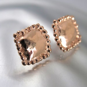 May include: A pair of rose gold square stud earrings. Each earring has a smooth, reflective center surrounded by a beaded border. The earrings are displayed on a reflective surface.