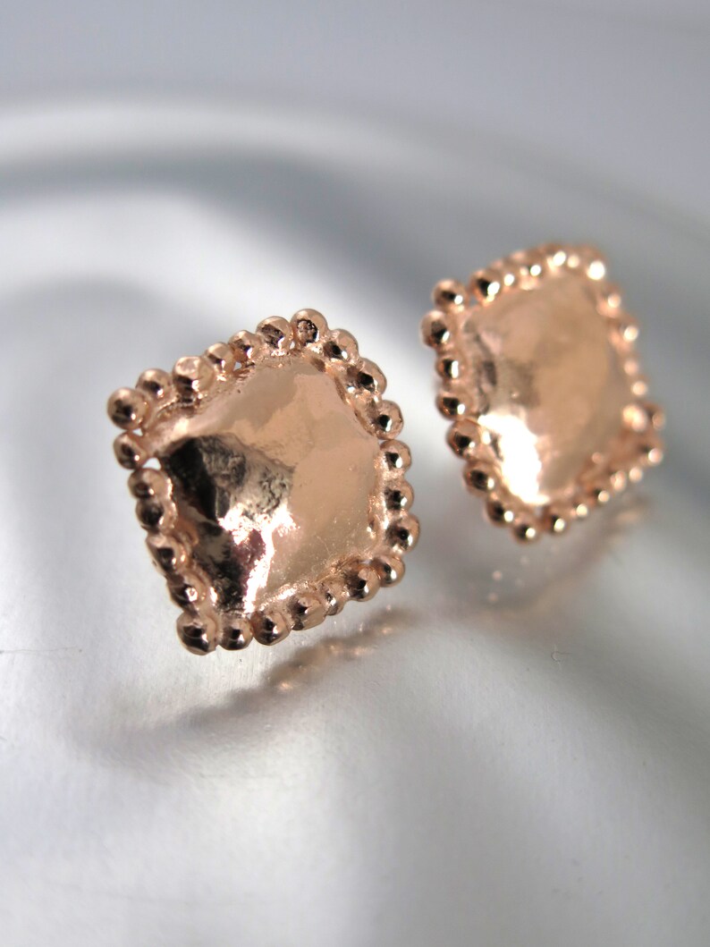 May include: A pair of rose gold square stud earrings. Each earring has a smooth, reflective center surrounded by a beaded border. The earrings are displayed on a reflective surface.