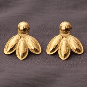 May include: A pair of gold-coloured earrings. Each earring has a round stud and three petal-shaped elements. The earrings are displayed on a grey background. The design is reminiscent of a stylized flower.