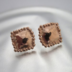 May include: A pair of rose gold square stud earrings. Each earring has a textured, square center surrounded by a beaded border. The earrings are displayed on a reflective surface.
