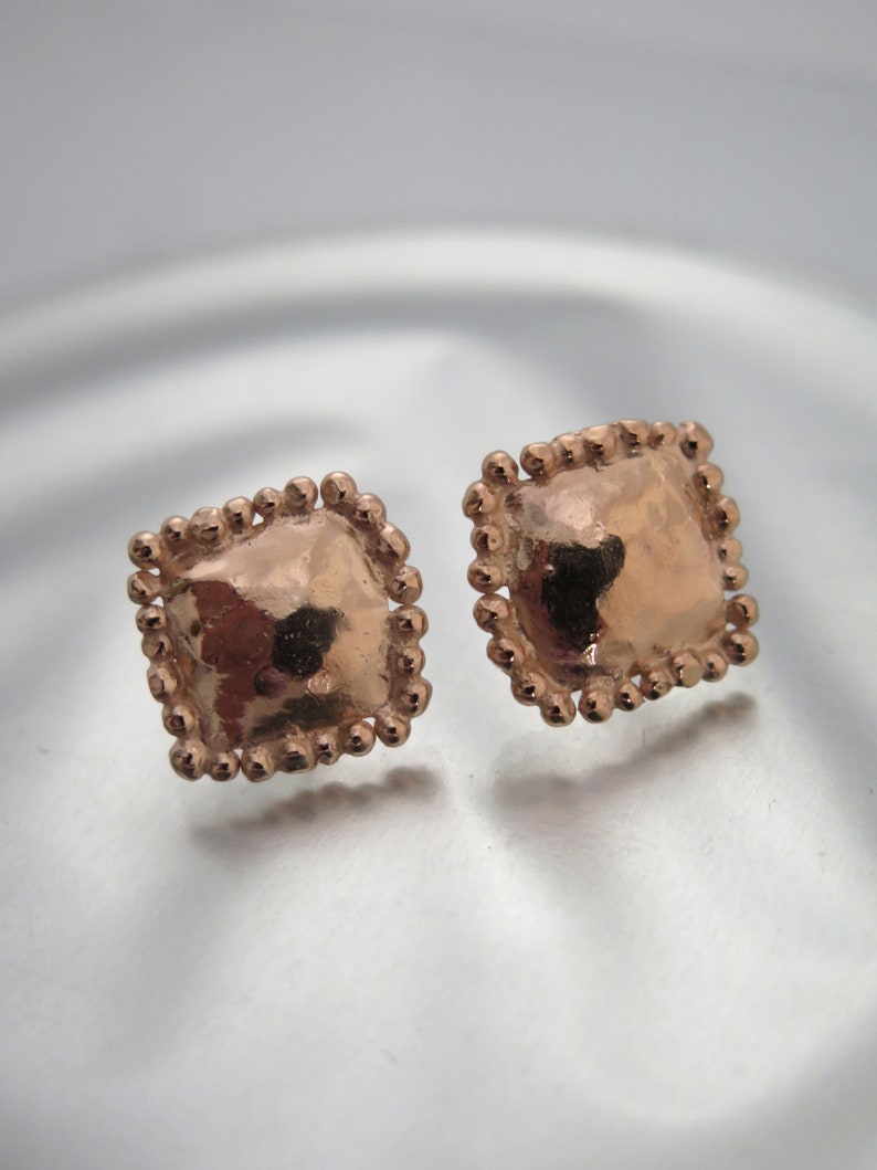 May include: A pair of rose gold square stud earrings. Each earring has a textured, square center surrounded by a beaded border. The earrings are displayed on a reflective surface.