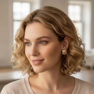 May include: A woman wearing gold square stud earrings. The earrings have a textured surface and a small, detailed design in the center. The woman has blonde, wavy hair and is wearing a light-colored top.