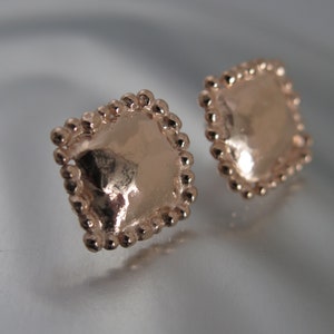 May include: Two rose gold square stud earrings. Each earring has a textured surface and is framed with small, round beads. The earrings are displayed on a reflective surface, with soft lighting.