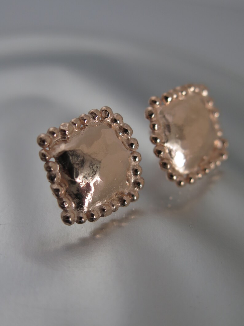 May include: Two rose gold square stud earrings. Each earring has a textured surface and is framed with small, round beads. The earrings are displayed on a reflective surface, with soft lighting.