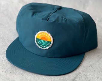 May include: A teal blue baseball cap with a patch that says "Vermont Lake Champlain" with a sunset and waves design.