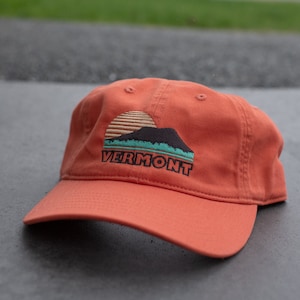 May include: An orange baseball cap with an embroidered design of a mountain range with a sun setting behind it. The text "VERMONT" is embroidered below the mountain range.