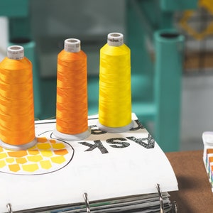 May include: Three spools of thread, one yellow and two orange, are sitting on a white page with the word "ASK" printed on it. The page is part of a binder with other pages visible.
