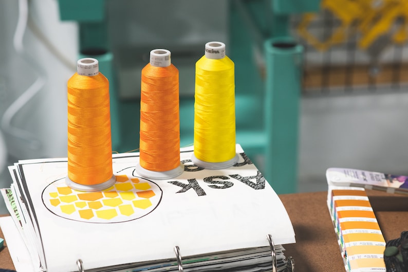 May include: Three spools of thread, one yellow and two orange, are sitting on a white page with the word "ASK" printed on it. The page is part of a binder with other pages visible.
