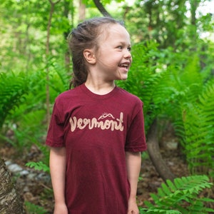 Puede incluir: Una persona joven lleva una camiseta granate con la palabra "Vermont" escrita en cursiva beige. La camiseta tiene un pequeño gráfico de montañas sobre la palabra. La persona está de pie en un bosque con helechos verdes.