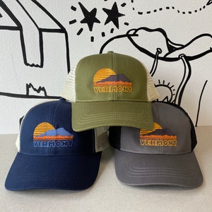 May include: Three trucker hats with a sunset and mountain graphic and the text "Vermont". The hats are in green, navy blue, and grey.
