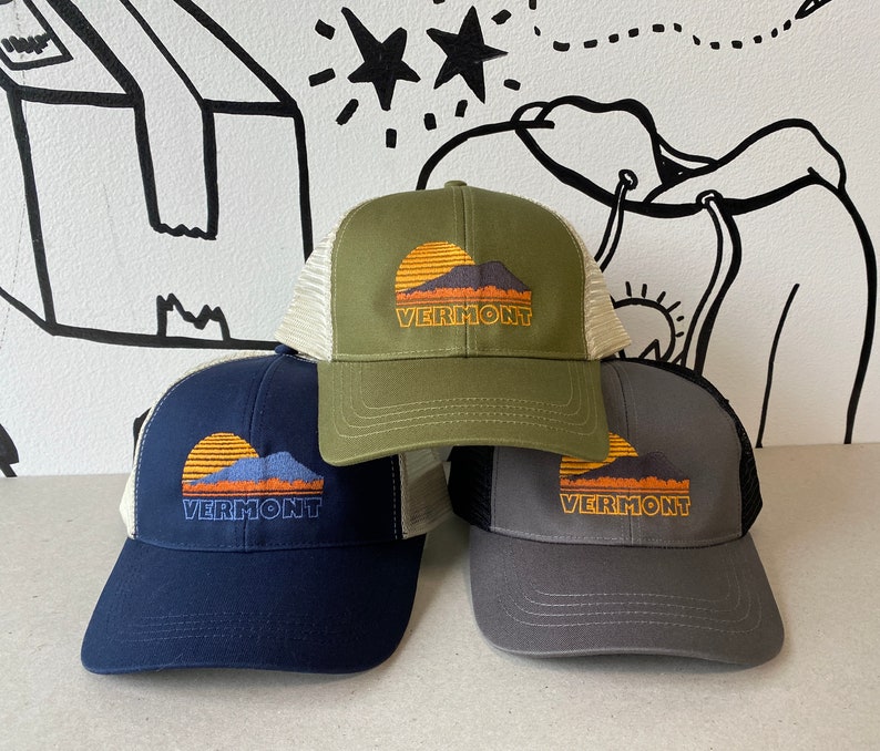 May include: Three trucker hats with a sunset and mountain graphic and the text "Vermont". The hats are in green, navy blue, and gray.