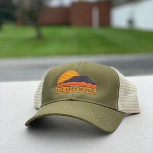 May include: A green and beige trucker hat with a sunset and mountain graphic and the word "VERMONT" embroidered on the front.