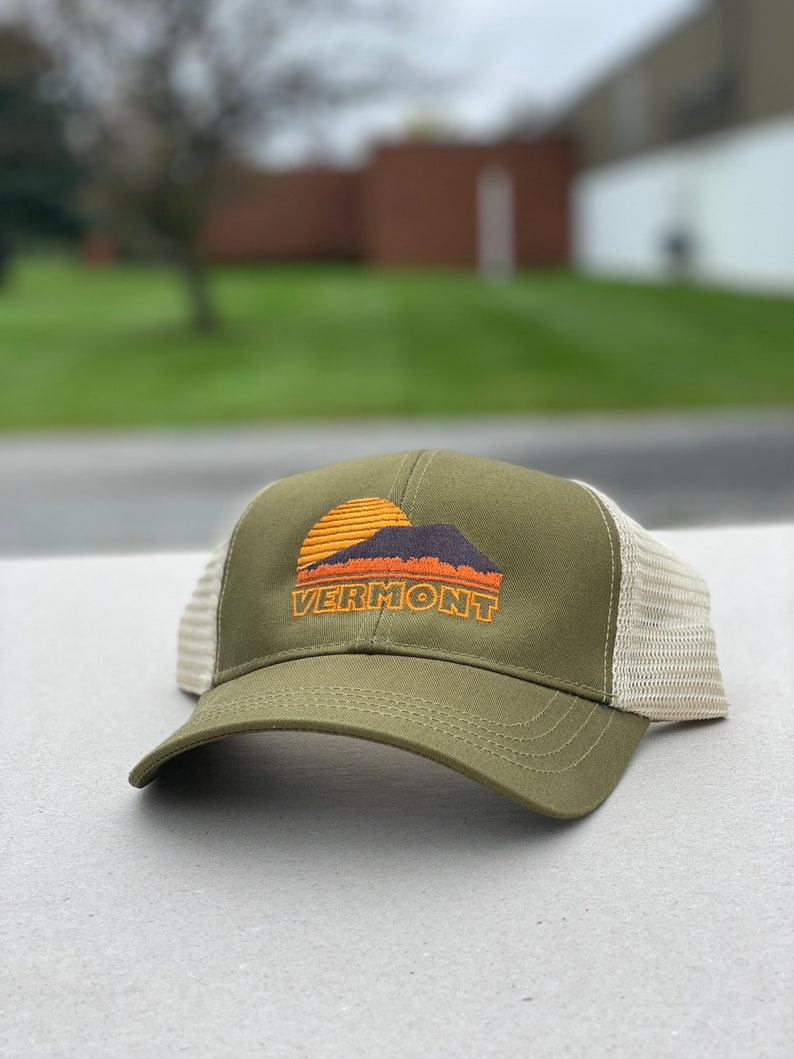 May include: A green and beige trucker hat with a sunset and mountain graphic and the word "VERMONT" embroidered on the front.