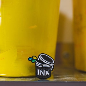 May include: Black enamel pin of an open paint can with colorful paint splatters and the word "INK" on the side.