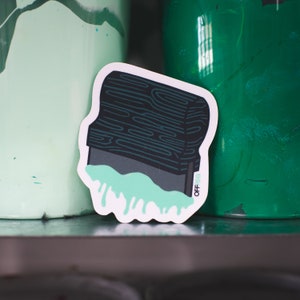 Op de afbeelding: Een sticker van een zwarte inktblok met een houtnerfstructuur, die groene verf laat druppelen. De sticker heeft de tekst "Off-Set" in wit.
