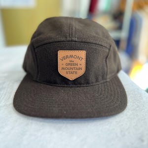 Puede incluir: Una gorra marrón de 5 paneles con un parche de cuero marrón que dice "Vermont the Green Mountain State".
