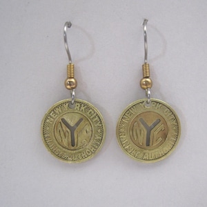 May include: A pair of gold-toned earrings with a circular design featuring the letters "NYC" in a cutout style. The earrings are suspended from silver-toned hooks.