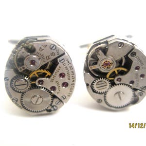 May include: A pair of cufflinks featuring the intricate mechanisms of vintage watch movements. The silver-toned cufflinks showcase gears, jewels, and other internal components. The text "SEVENTEEN 17 JEWELS" is visible on the watch parts. The date "14/12/2014" is also visible.