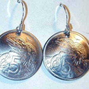 May include: Silver-toned coin earrings. Each earring features a round coin with a detailed lizard design and the number 5. The earrings have silver-colored hooks.
