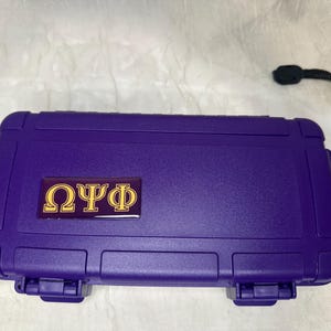 Könnte beinhalten: Ein lila rechteckiger Koffer mit einem schwarzen Schlüsselband. Der Koffer hat ein gold-violettes Emblem mit griechischen Buchstaben. Der Koffer hat eine strukturierte Oberfläche und einen sicheren Verschluss.