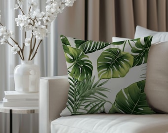 May include: Square throw pillow with a tropical leaf pattern. The pillow features large green monstera leaves and other tropical foliage on a white background. The pillow is placed on a white sofa.