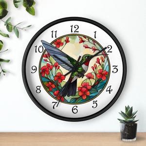 May include: Round wall clock with a black frame and a white face. The clock features a colorful illustration of a hummingbird with red flowers. The clock hands are black, and the numbers are in a classic font.