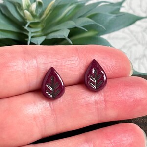 May include: A pair of teardrop-shaped stud earrings in a deep burgundy colour. Each earring features a glossy finish and a recessed, dark-coloured leaf pattern etched into the centre of the surface, creating a distinct contrast against the smooth base.