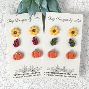 May include: Two white display cards labelled Clay Designs by Mia, each holding three pairs of stud earrings. The earrings feature yellow sunflowers with brown centres, maroon or olive green leaves, and orange pumpkins with metallic gold stems.