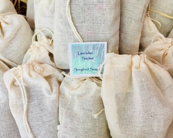 May include: A collection of off-white fabric sachets filled with lavender, tied with a drawstring. A small card reads "Lavender Sachet" and "Songbird Soap" against a blue and green watercolor background. The sachets are arranged in a woven basket.
