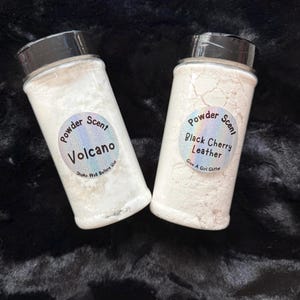 Könnte beinhalten: Zwei durchsichtige Kunststoff-Streudosen gefüllt mit weißem Pulver, jeweils mit einem schwarzen Deckel versehen. Die linke Dose hat ein holografisches rundes Etikett mit der Aufschrift Powder Scent Volcano Shake Well Before Use. Das rechte Etikett zeigt Powder Scent Black Cherry Leather Give A Girl Glitter.