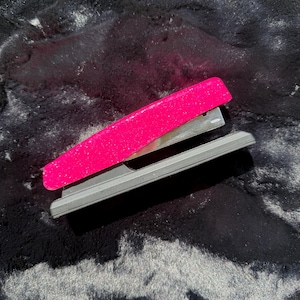 May include: A hot pink stapler with silver metal components and a gray base. The stapler is set against a dark, textured background.