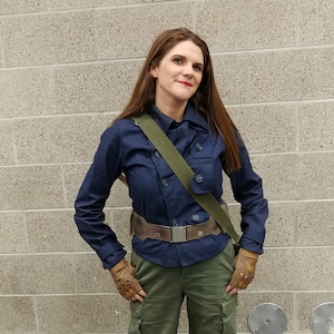 May include: A dark blue button-up jacket with a collar, olive green cargo pants with side pockets, a brown utility belt with a silver buckle, a green shoulder strap, tan fingerless gloves, and black lace-up boots reaching mid-calf.