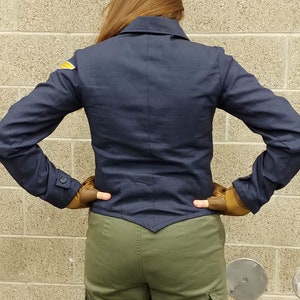May include: A dark navy blue jacket with a pointed hem, a collar, and long sleeves featuring buttoned cuffs. A small yellow and white wing-shaped patch is visible on the right shoulder. The jacket is paired with olive green cargo pants.