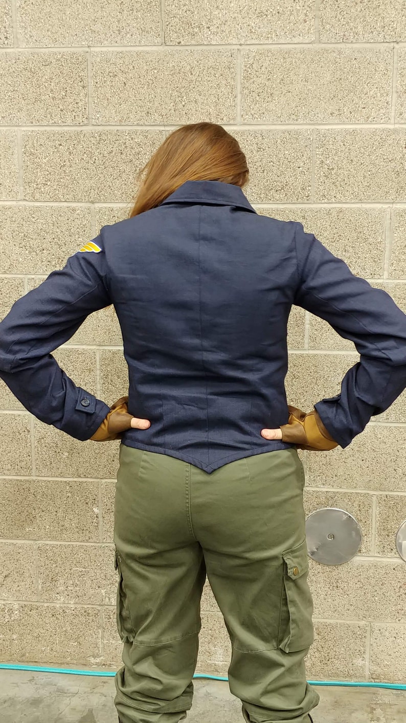 May include: A dark navy blue jacket with a pointed hem, a collar, and long sleeves featuring buttoned cuffs. A small yellow and white wing-shaped patch is visible on the right shoulder. The jacket is paired with olive green cargo pants.