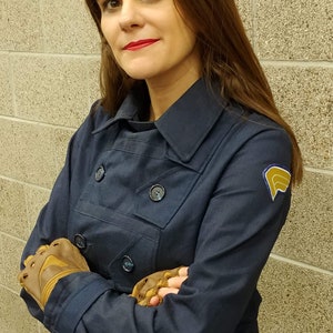 May include: A dark navy blue double-breasted jacket with a structured collar, long sleeves, and dark buttons. The jacket features a yellow and blue embroidered patch on the left shoulder. The garment is paired with brown fingerless leather gloves.